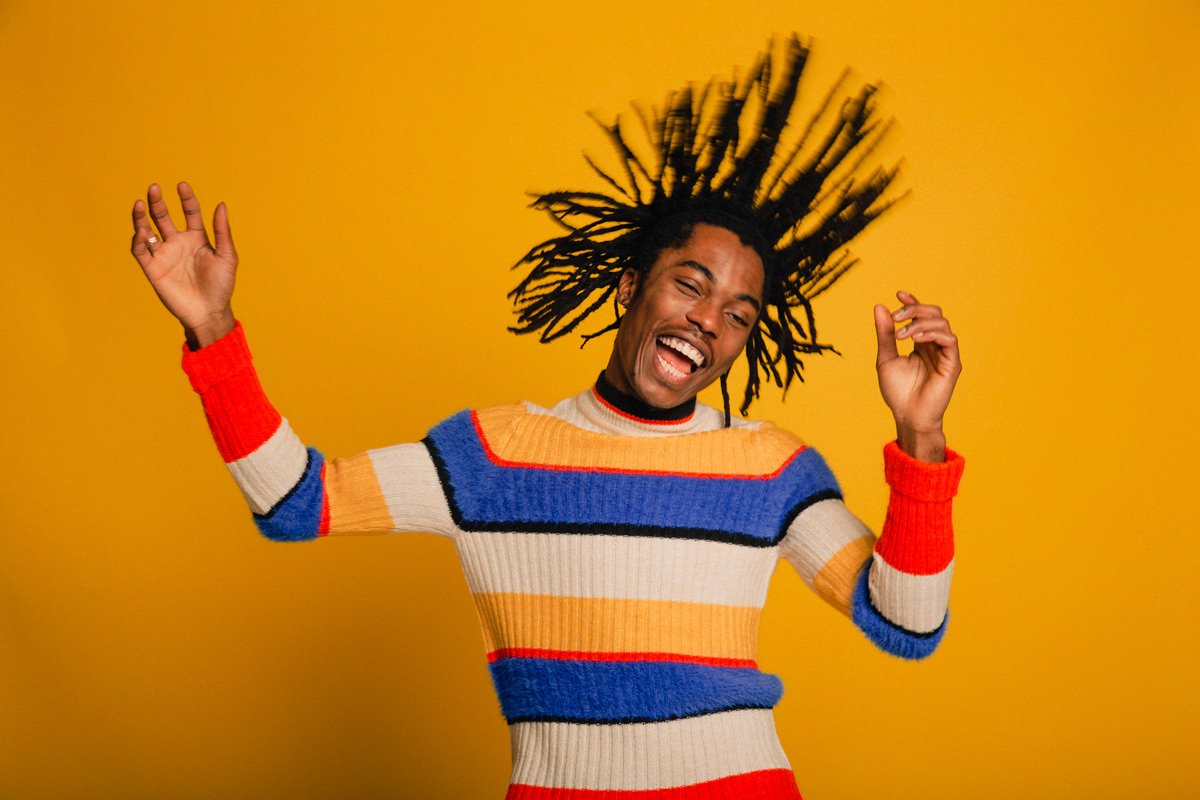 A man with dread locks laughs as he flips his hair back and forth in a brightly colored sweater against a yellow background.