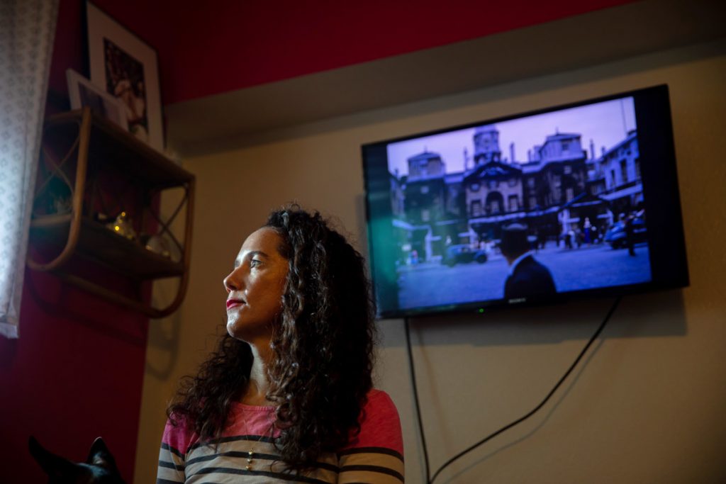 A woman looks off into the distance toward a window with an old photograph on a television screen behind her. 