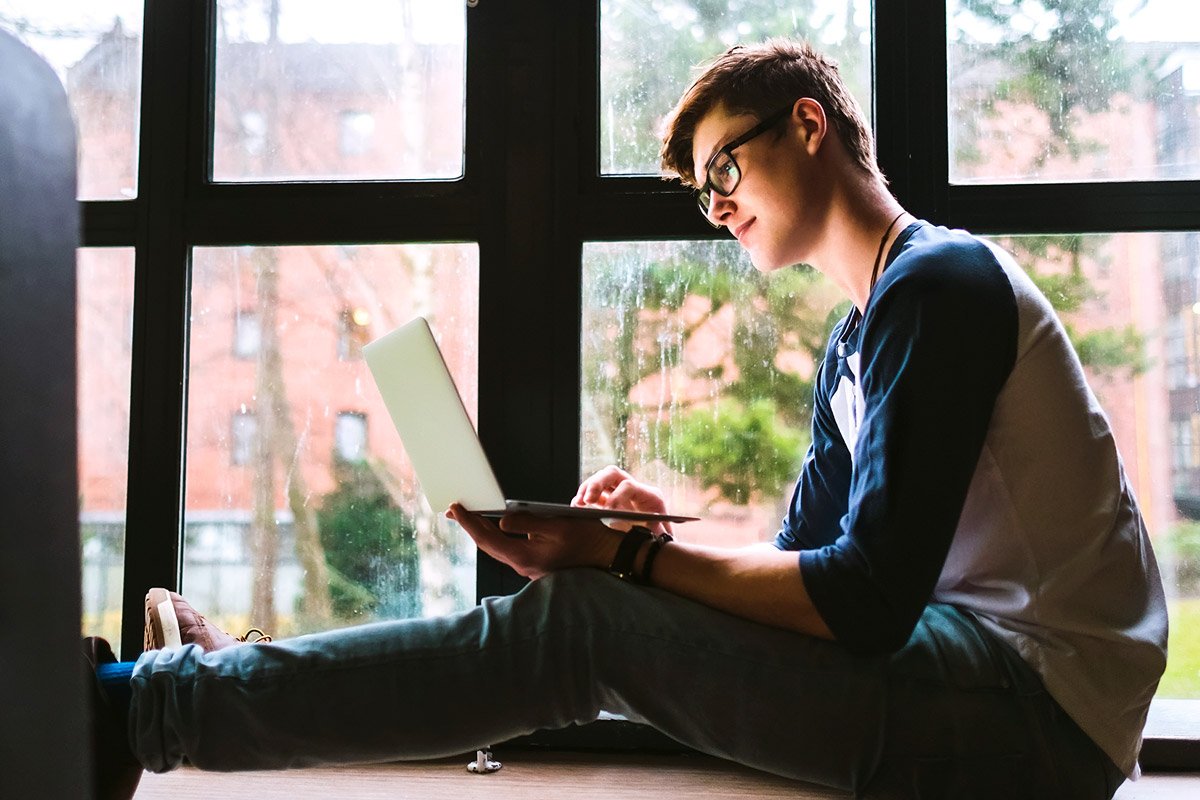 A college student sits next to a window while working on his laptop. 