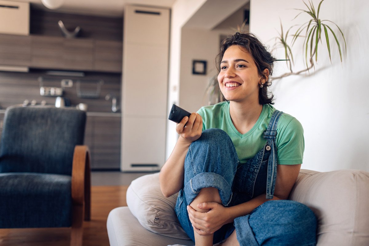 A woman smiles as she holds her remote in her hand while watching television from the comfort of her couch.