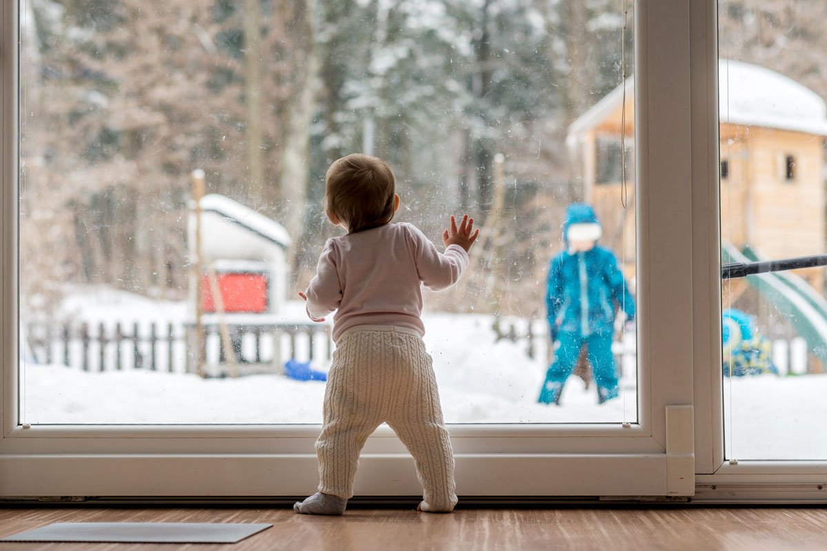 A toddler watches his sibling play out in the snow from the window. 