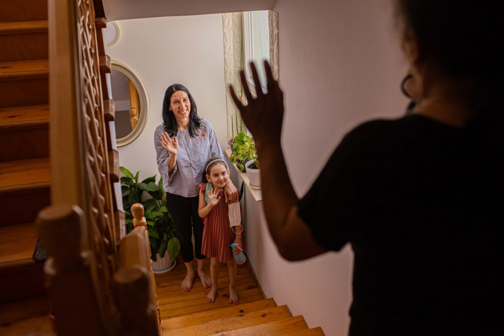 A little girl waves to a person while standing next to her babysitter.