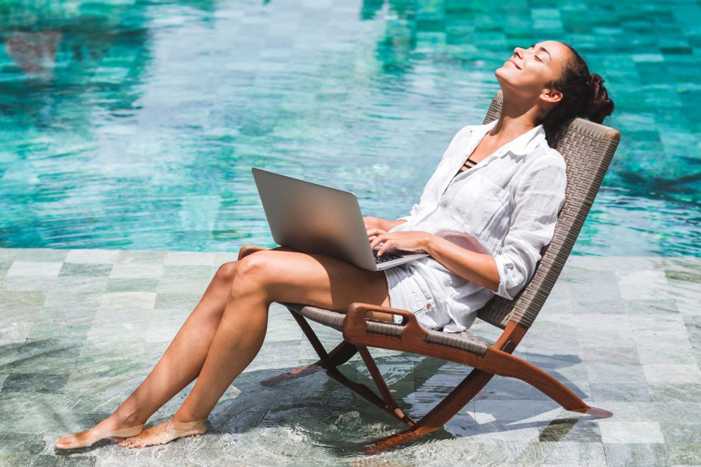 A woman works on her laptop while dipping her feet into a pool.