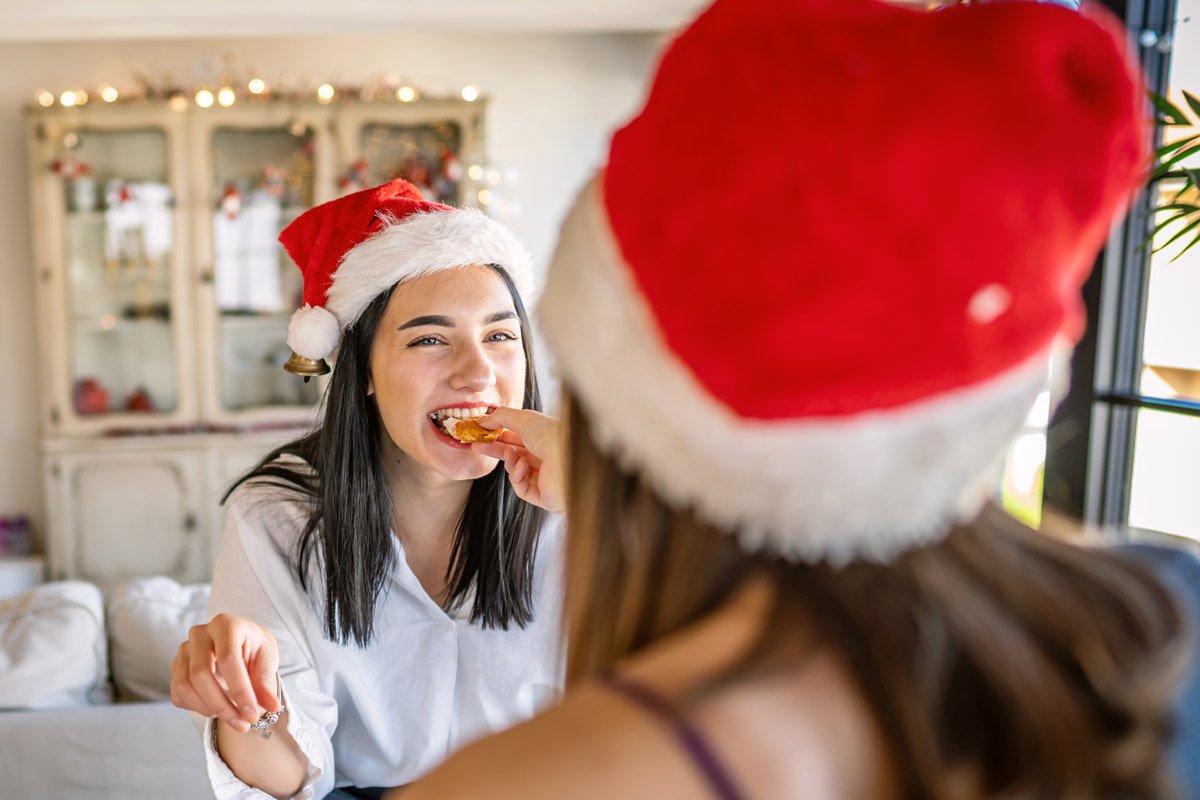 A lesbian couple feed each other while wearing Santa hats. 