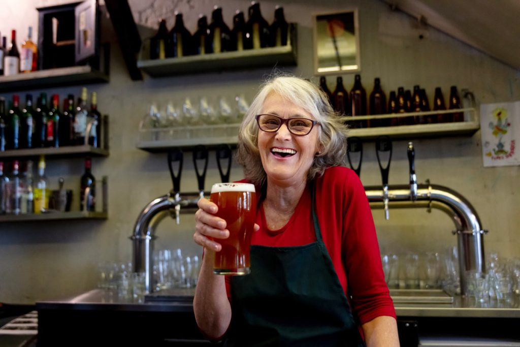 A senior citizen bartender holds up a pint of beer. 