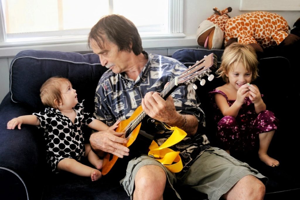 An elderly man babysits two girls. He plays guitar on the couch while the two of them listen to him play.
