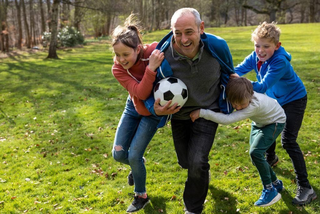 A group of kids chase after their grandfather with the soccer ball.