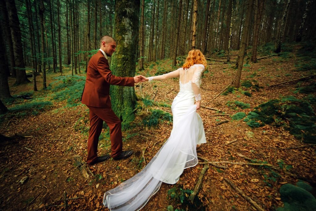 A bride and groom walk hand in hand in a forest. 