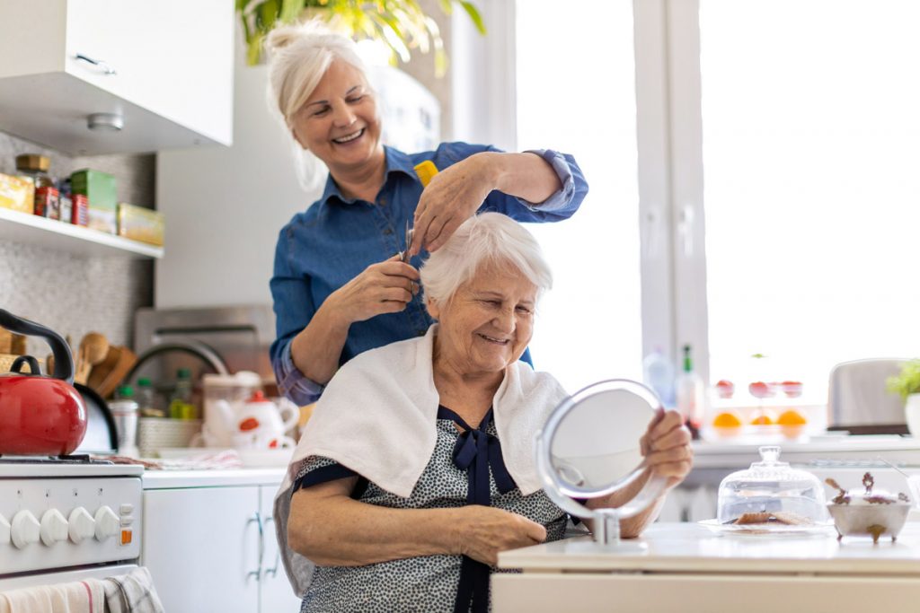 A woman cuts an elderly woman's hair at her kitchen table.