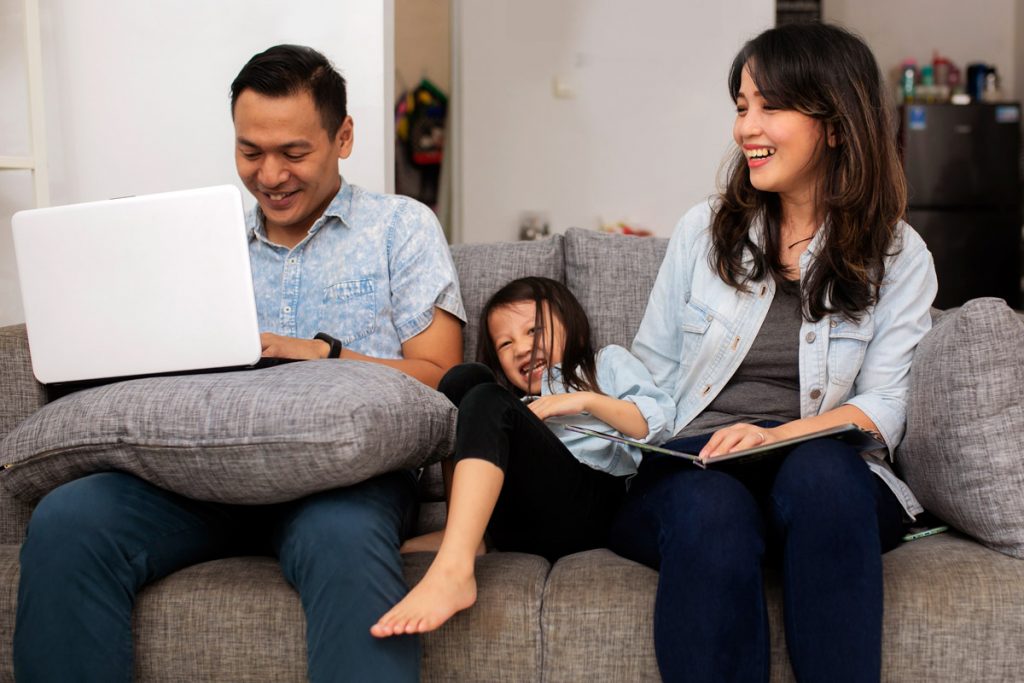 A man and wife laugh at their daughter as the man does work on his laptop on their couch.