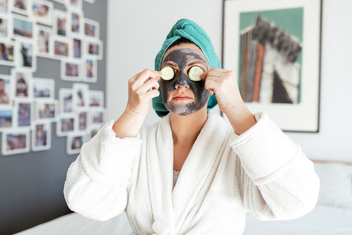 A woman puts cucumbers over her eyes as she sits up with a charcoal facial mask on her. 