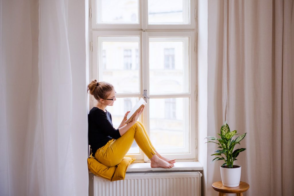 A woman sits on a windowsill while reading a book.