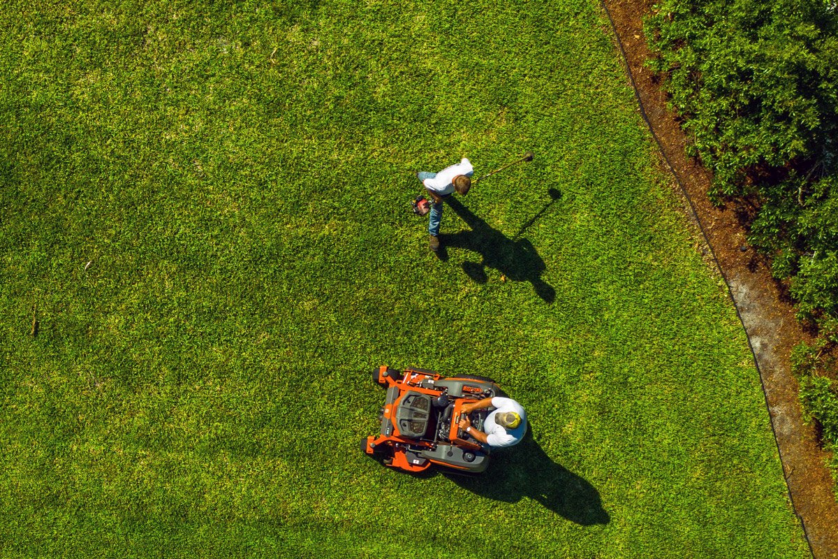 Two people work on a person's yard by mowing it and trimming bush hedges.