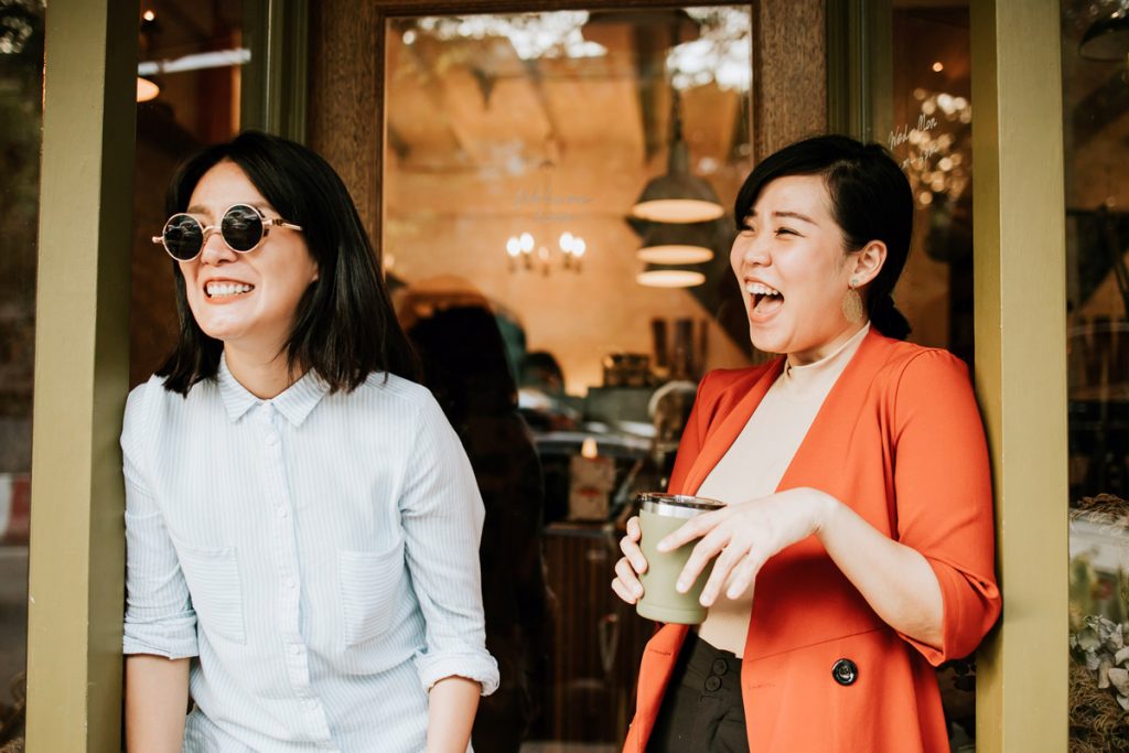 Two women laugh outside of a cafe while dressed in professional work clothes.