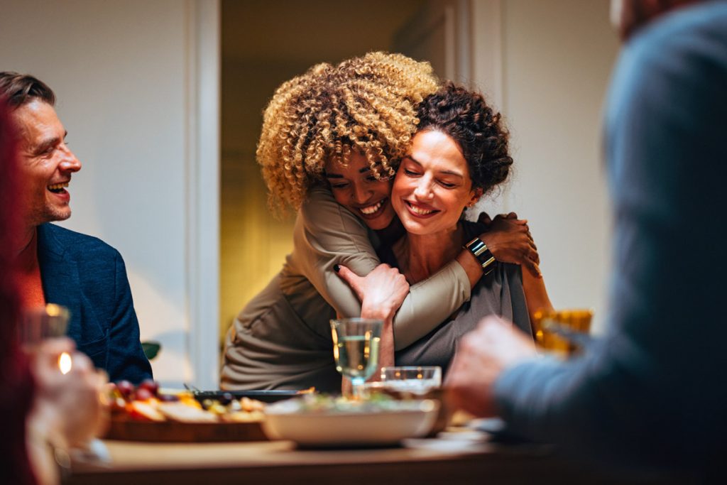 A group of friends host a dinner party where two of them hug at the dinner table.
