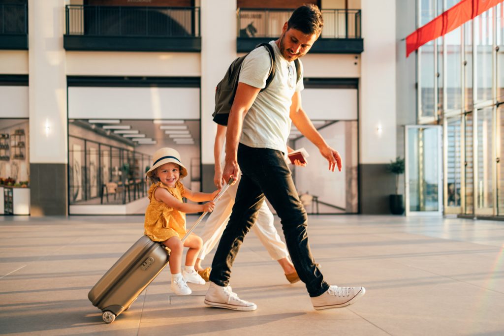 A man walks through the airport with her daughter riding on his luggage.