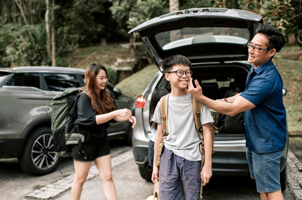 A father jokingly touches his son's cheek as they pack their vehicle for a road trip.
